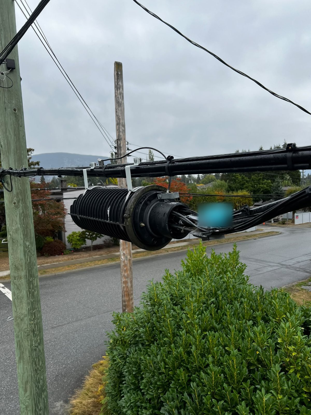 Aerial fiber splice closure mounted on the strand above a residential street, with a cable tag obscured